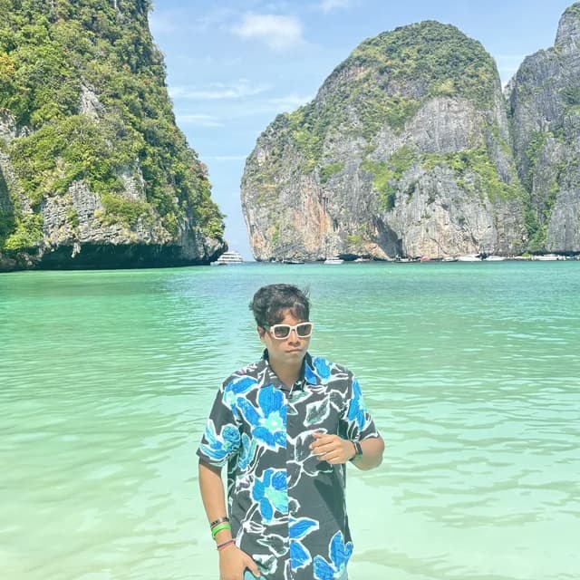 Adarsh Soni, in sunglasses and a blue floral shirt, standing in clear turquoise water below limestone cliffs